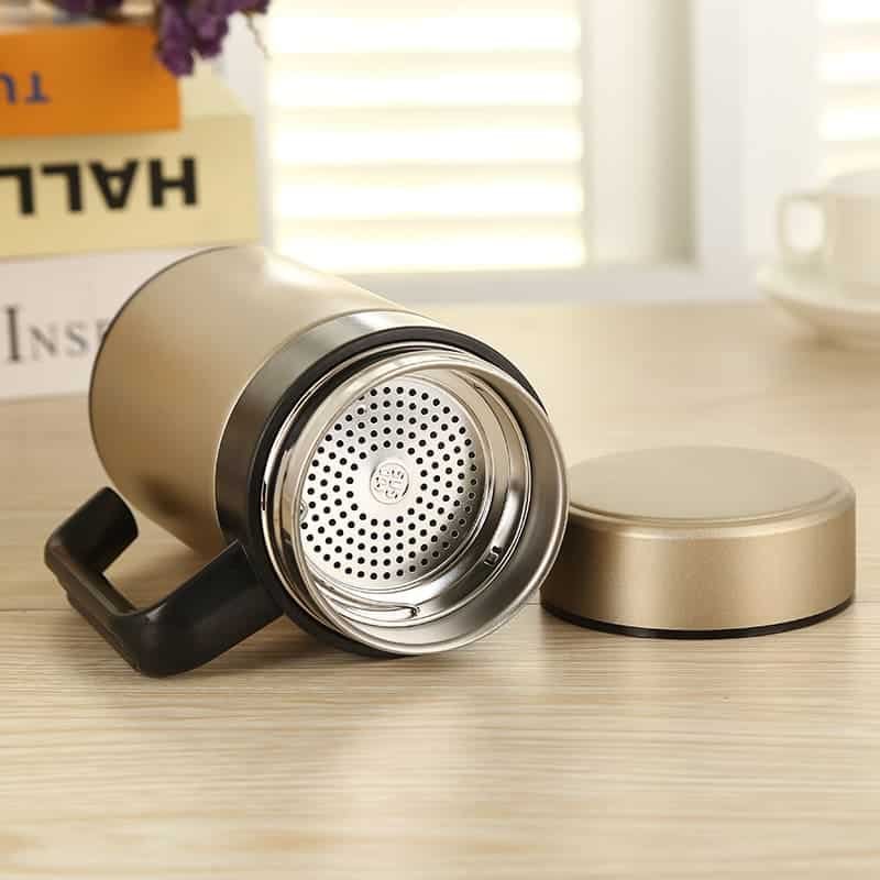 An angled top-down interior view of a gold office mug revealing the clean SUS 304 metal stamp and a detached tea filter.