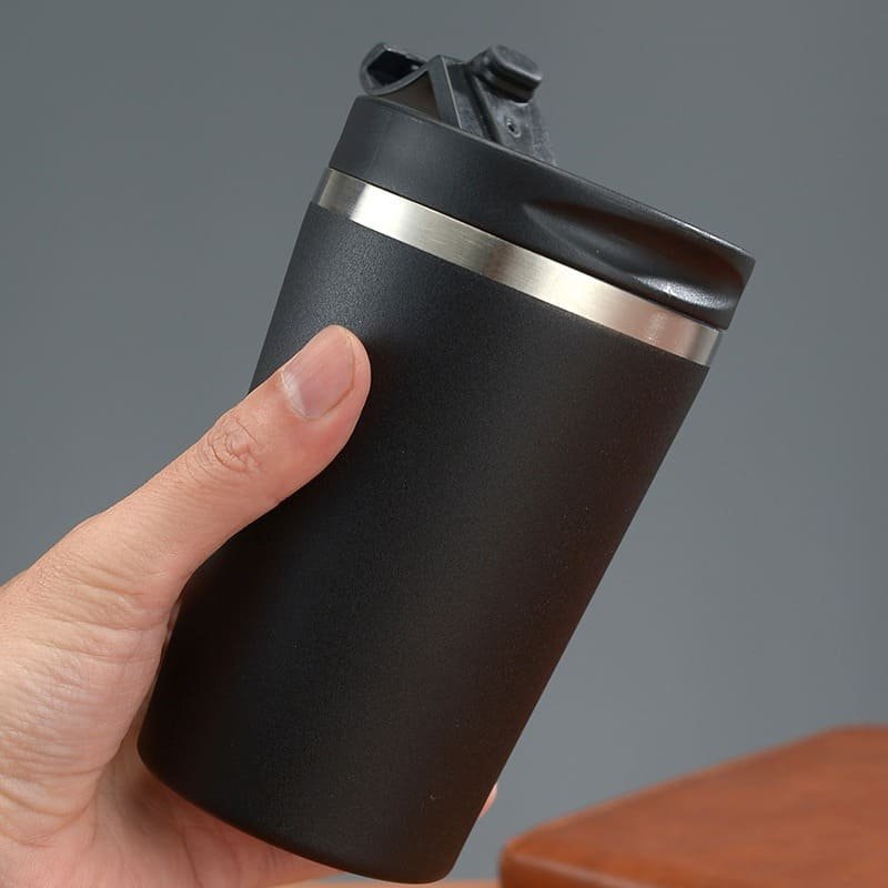 A hand holding the matte black 320ml vacuum coffee mug with the snap top flip lid partially opened against a gray background.
